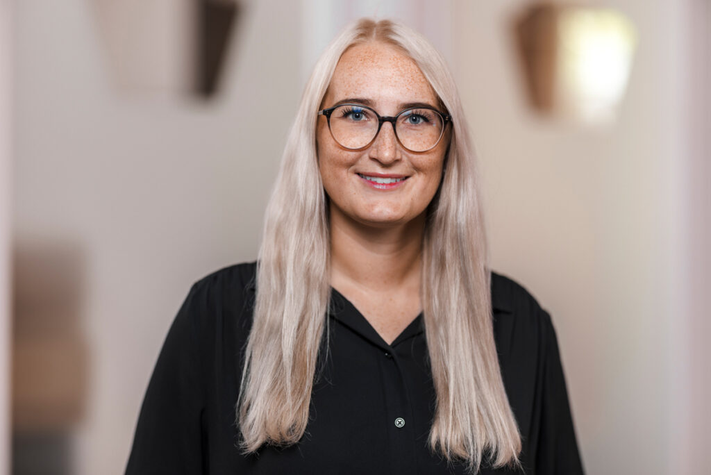 Porträt einer jungen Frau mit langen blonden Haaren und Brille, sie trägt eine schwarze Bluse und lächelt leicht in die Kamera, neutraler Hintergrund, professionelles Foto für Unternehmens- oder Teamprofil.