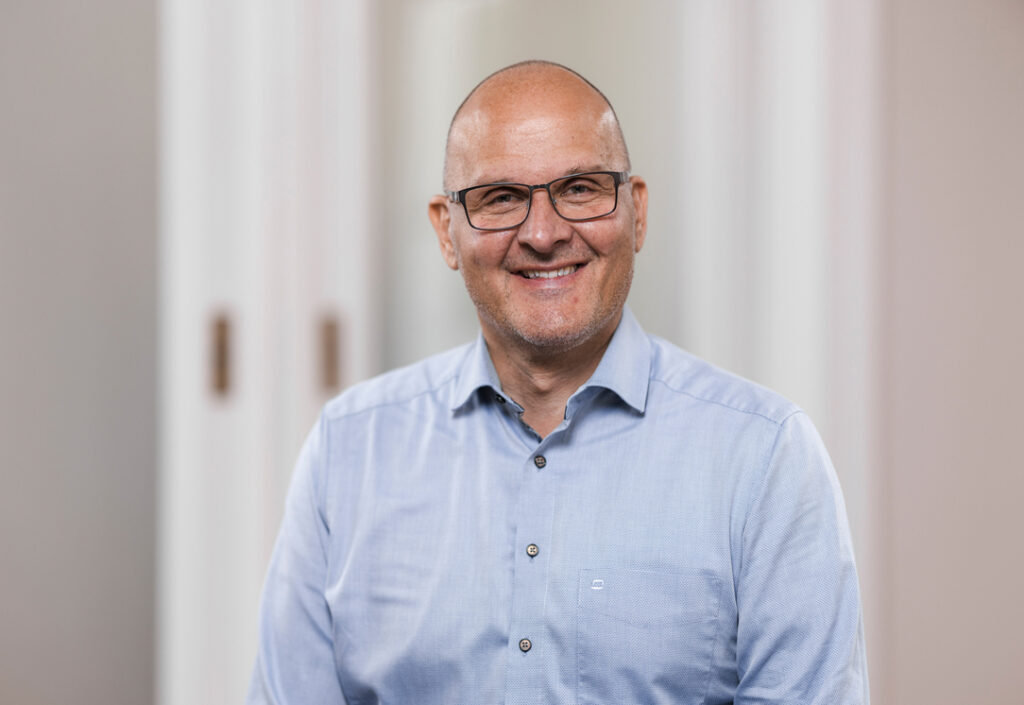 Porträt eines lächelnden Mannes mit Brille und hellem Hemd, Glatze, freundlicher Gesichtsausdruck, neutrale Hintergrundumgebung, professionelles Foto für Unternehmens- oder Teamprofil.