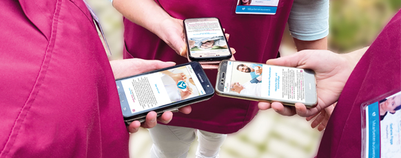 Mehrere Pflegekräfte in bordeauxfarbenen Kasacks halten ihre Smartphones zusammen, auf den Bildschirmen sind digitale Informationen und Pflege-Apps zu sehen – Symbol für moderne Kommunikation und digitale Pflegeorganisation.