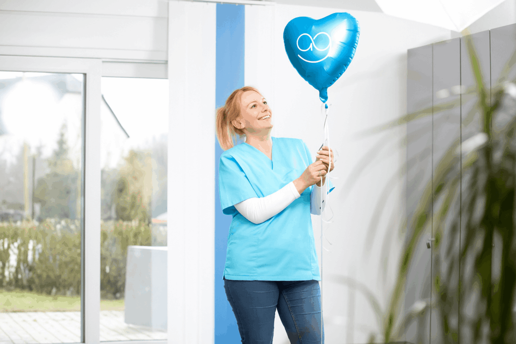 Junge blonde Pflegekraft in hellblauer Arbeitskleidung steht lächelnd in einem hellen Raum und hält einen blauen Herzballon mit Smiley-Motiv.