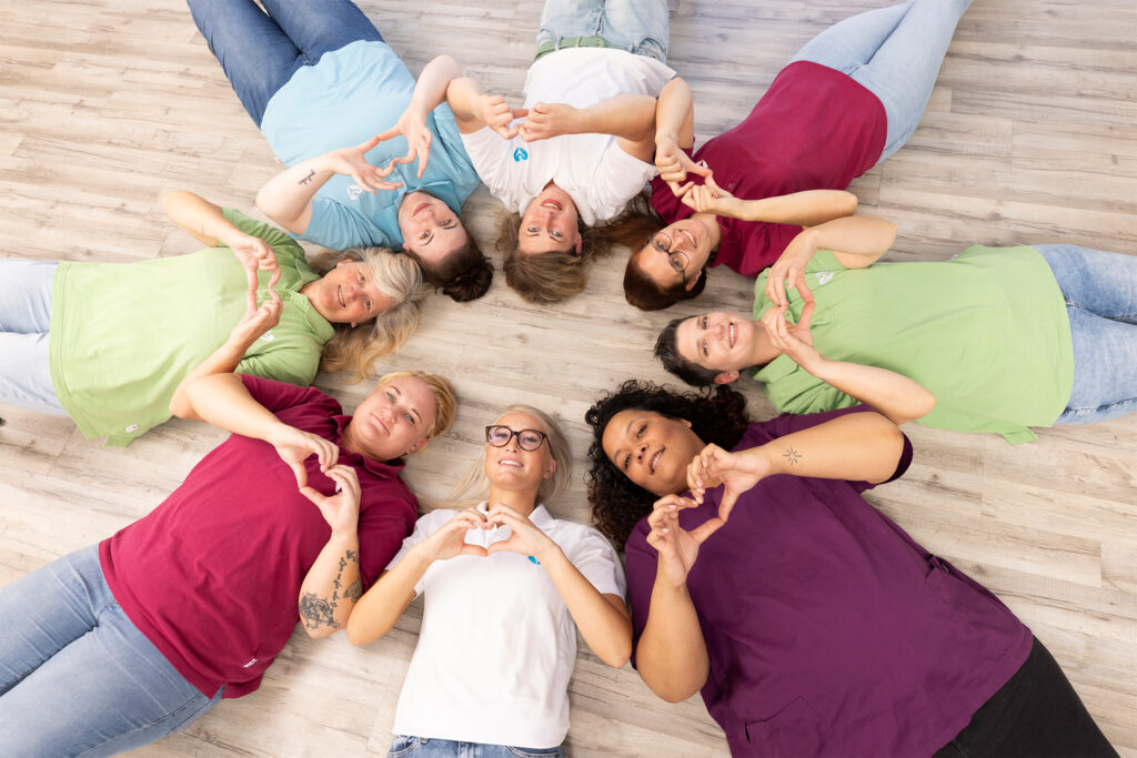 Pflegeteam liegt im Kreis auf dem Boden und formt mit den Händen Herz-Symbole. Acht Mitarbeiterinnen in bunten Polo-Shirts (grün, weiß, blau, bordeaux und lila) lächeln in die Kamera. Symbol für Teamgeist, Zusammenhalt und Wertschätzung in der ambulanten Pflege.
